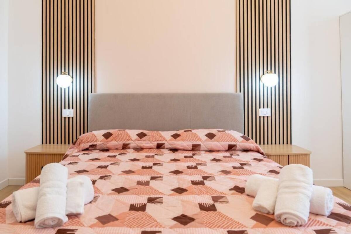 Un lit bien fait, recouvert d’une courtepointe à motifs, deux tables de chevet et deux lampes fixées au mur.