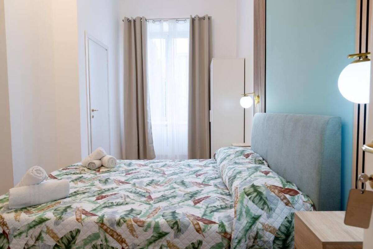 Une chambre d’hôtel avec un lit, une chaise, une table de chevet, une lampe et une fenêtre avec des rideaux.