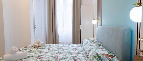 Une chambre d’hôtel avec un lit, une chaise, une table de chevet, une lampe et une fenêtre avec des rideaux.