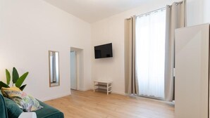 Un salon moderne avec un canapé vert, un téléviseur à écran plat fixé au mur, une petite table blanche et une grande fenêtre avec des rideaux.