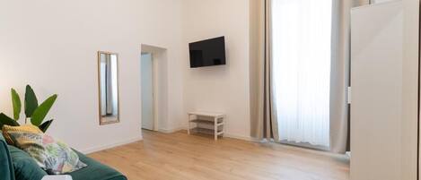 Una sala moderna con un sofá verde, un televisor de pantalla plana montado en la pared, una mesita blanca y un ventanal con cortinas.