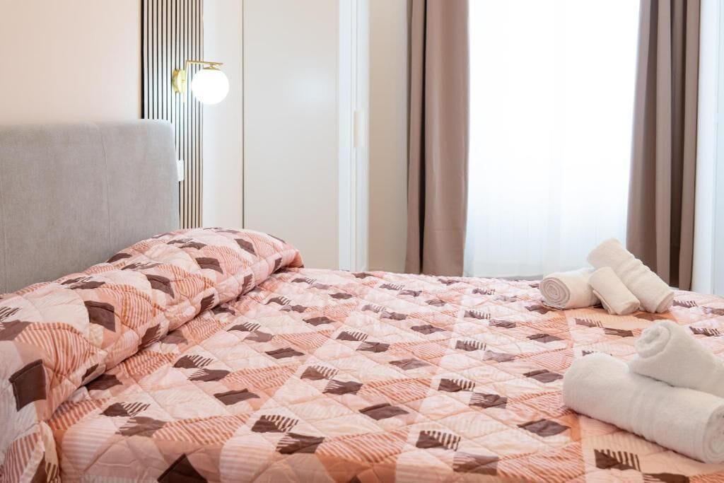 Una cama bien tendida con un edredón estampado, una lámpara de noche y toallas enrolladas en el suelo.