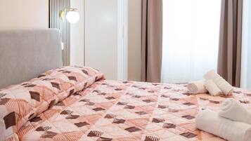 Una cama bien tendida con un edredón estampado, una lámpara de noche y toallas enrolladas en el suelo.