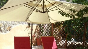 Deux fauteuils de jardin en bois, dotés de coussins rouges, sont placés sous un parasol beige, sur une terrasse en pierre bordée de gravier.