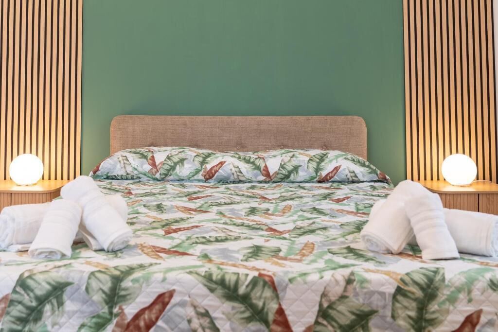A neatly made bed with a tropical leaf pattern bedspread, two bedside lamps, and a green wall.