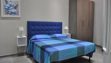 Ein Schlafzimmer mit blau gestepptem Kopfteil, einer karierten Tagesdecke, zwei Nachttischen mit Lampen und einem Kleiderschrank.