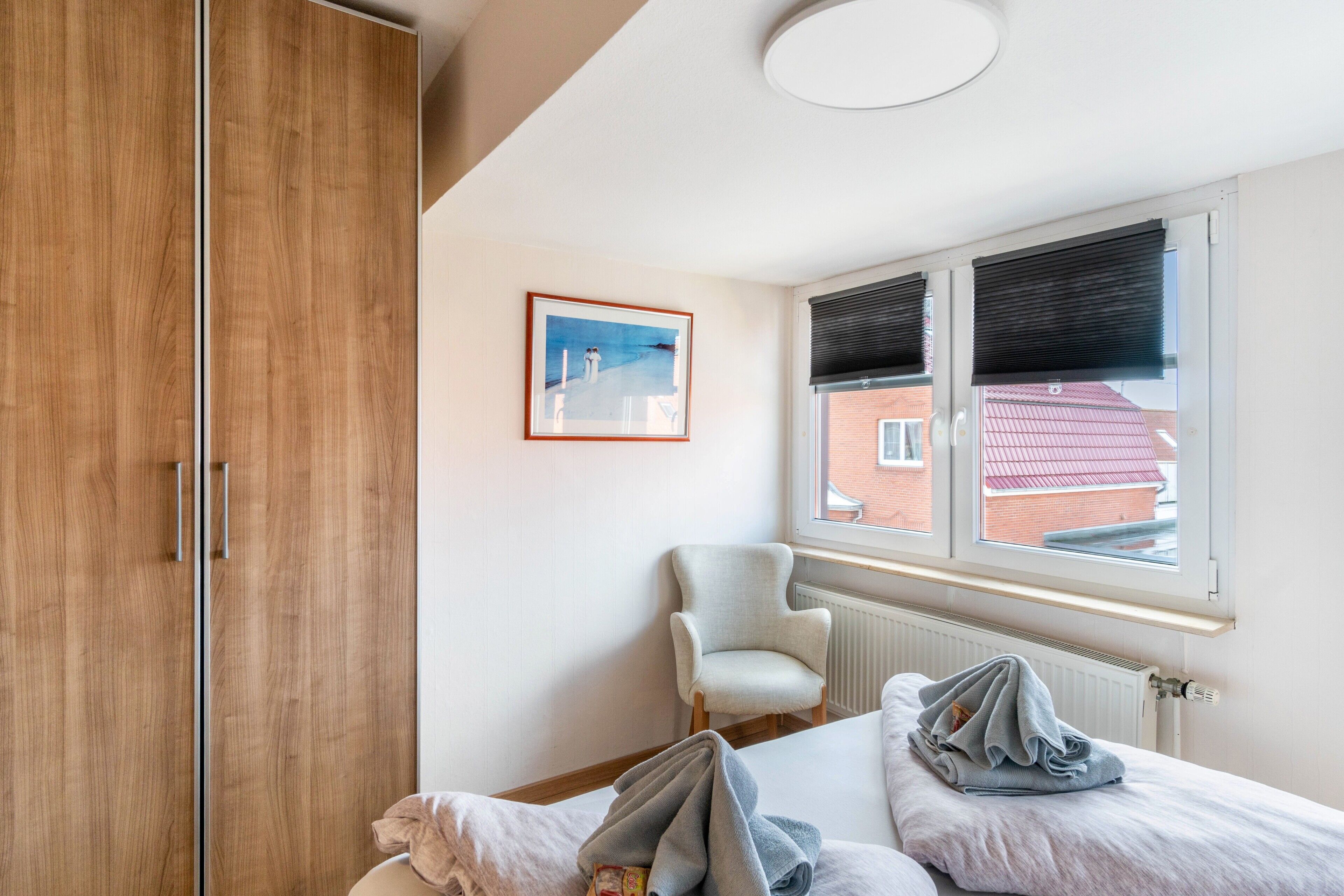 A bedroom with a wooden wardrobe, a single bed, a chair, a window with blinds, and a framed picture on the wall.