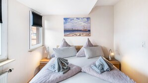 A bedroom with a bed, pillows, a painting of a beach scene, a window, and a nightstand with a lamp. - Apartment "Inselstern" on Borkum – 2 Bedrooms, Modern Kitchen, Wi-Fi (Borkum)