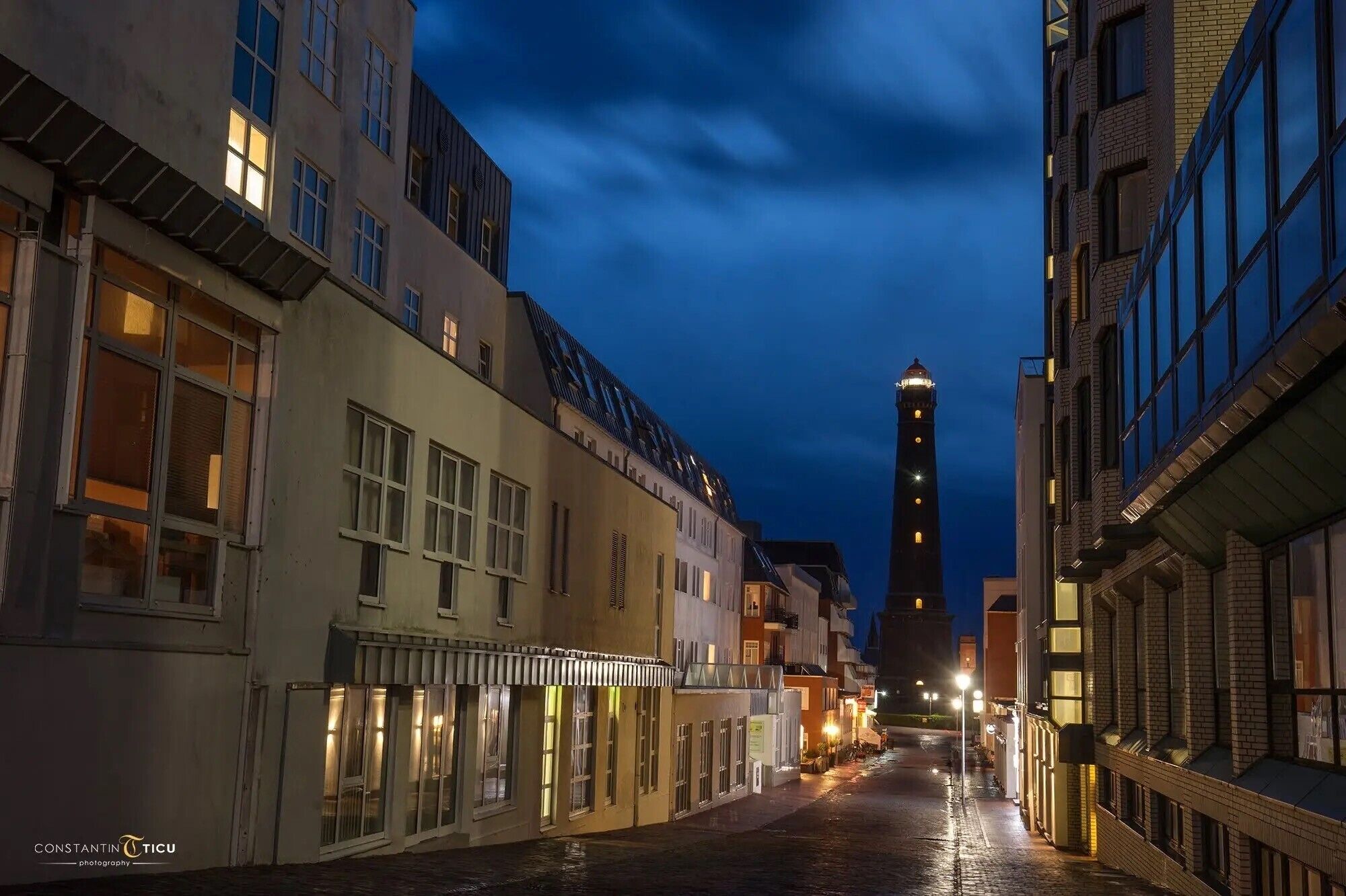 Eine beleuchtete Straße bei Nacht mit Gebäuden, einem markanten Turm und nasser Fahrbahn.