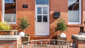 A brick building with a white door, two potted plants, a mailbox, and a small outdoor seating area. - Holiday Home 'Südstrand' on Borkum for 2–4 Guests, 2 Bedrooms (Borkum)