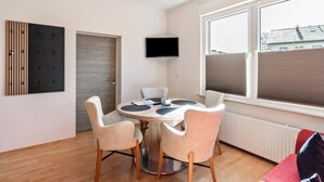 A modern room with a round dining table, four chairs, a flat-screen TV, and large windows. - Holiday Home 'Südstrand' on Borkum for 2–4 Guests, 2 Bedrooms (Borkum)
