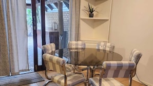 A dining area with a glass table and striped cushioned chairs, a white shelf with a plant, and a view through a glass door. - 5 Beach Dunes (Richards Bay)