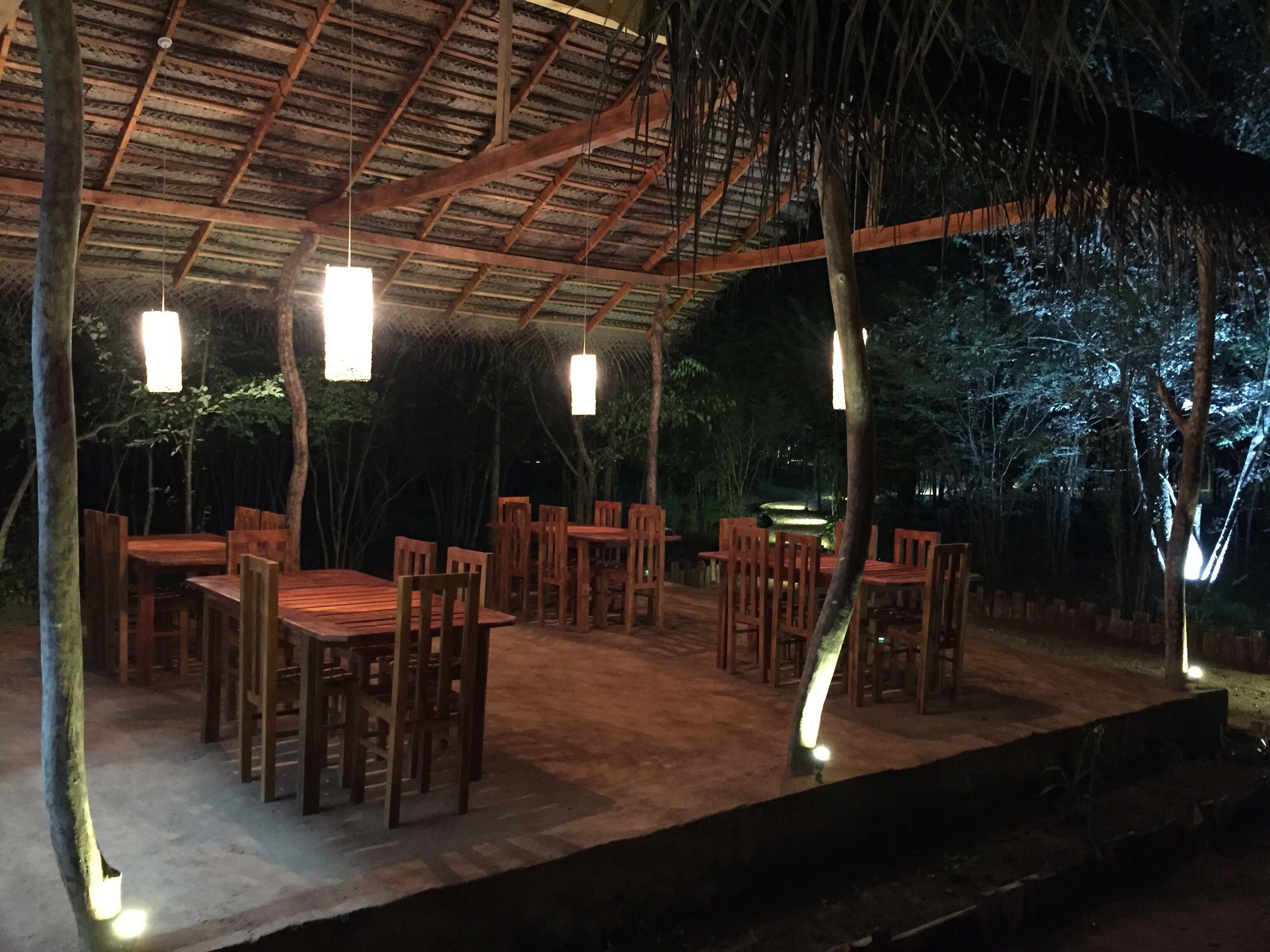 Un área de comedor al aire libre con mesas y sillas de madera, bajo un techo de paja con luces colgantes.