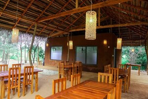 An outdoor dining area with wooden tables and chairs, hanging light fixtures, and a thatched roof structure.