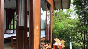 A wooden balcony with wicker chairs and a small table, overlooking a lush green area.