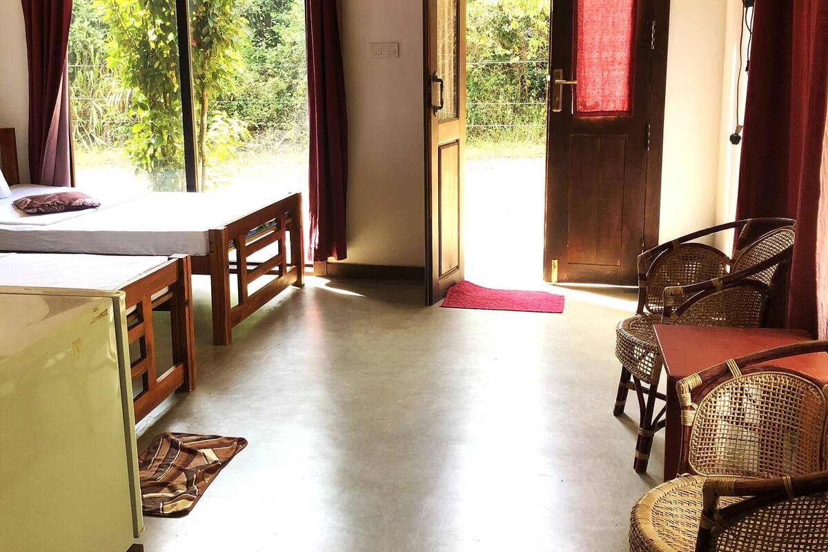 Quarto com cama, mesa, cadeiras, porta e vista para o verde lá fora.
