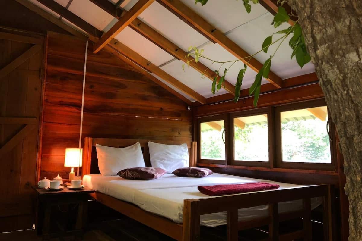 Quarto de cabana de madeira com cama, almofadas, mesa de cabeceira, abajur e janela.