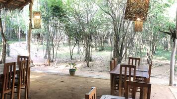 An outdoor dining area with wooden tables and chairs, a thatched roof, and a view of trees.