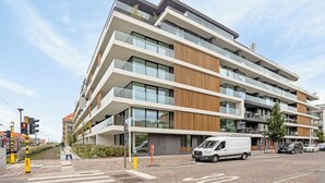 A multi-story modern building with large windows and balconies, a white van parked in front, and a pedestrian crossing nearby. - The Green 15 0301 met garage (Nieuwpoort)