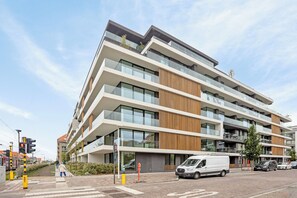 Ein mehrstöckiges, modernes Gebäude mit großen Fenstern und Balkonen, ein weißer Transporter vor dem Haus und eine Fußgängerüberweg in der Nähe.