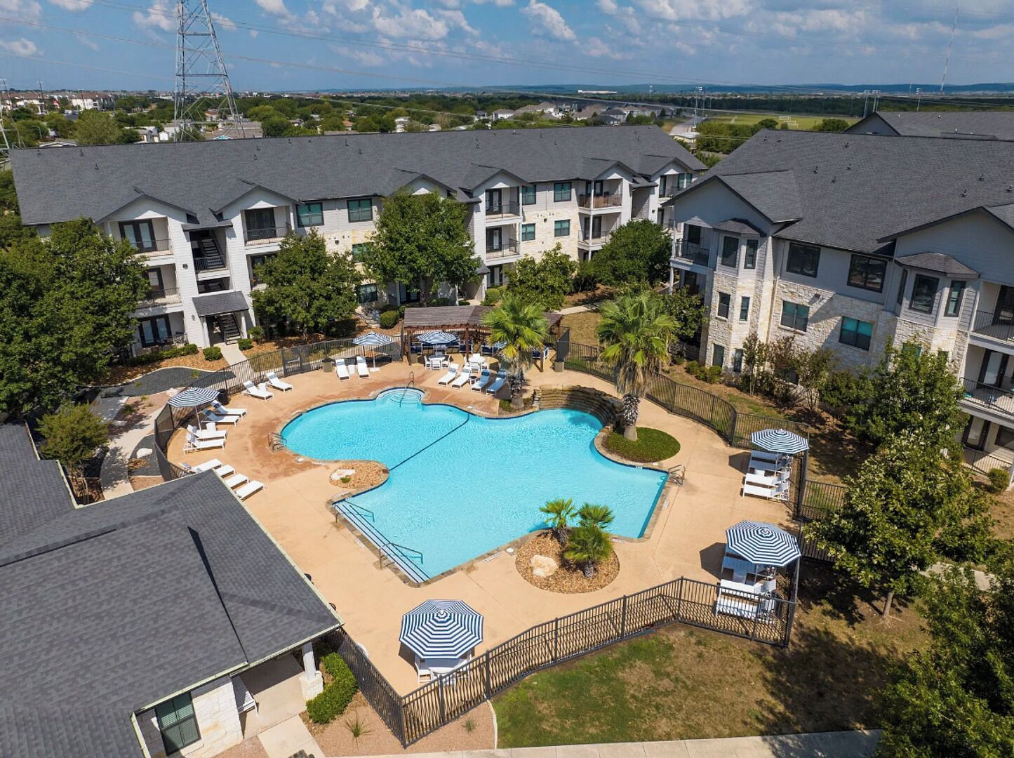 A residential complex with a central swimming pool surrounded by lounge chairs and umbrellas, set in a landscaped area with trees and a view of the surrounding landscape.