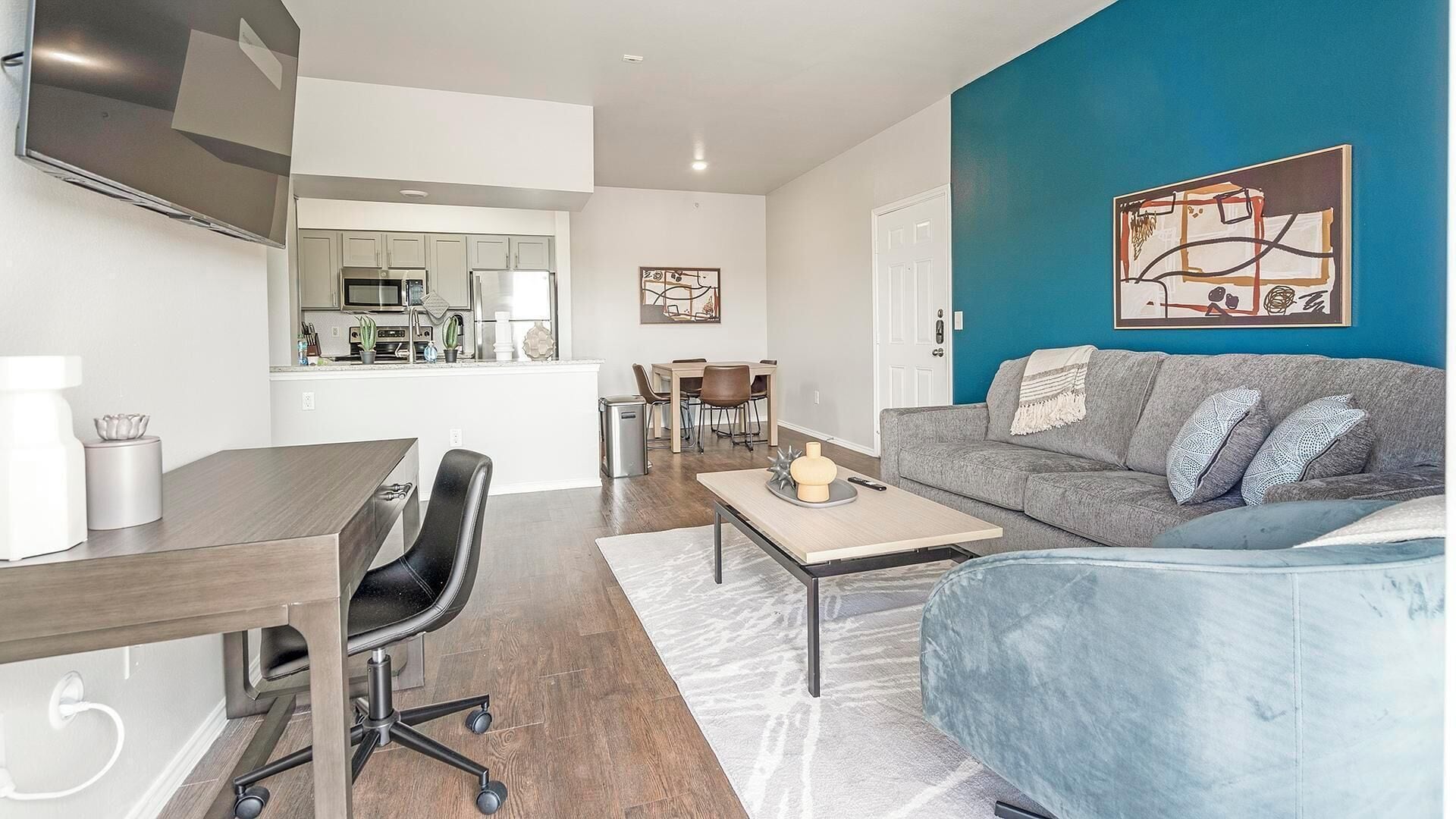 A modern living room with a grey sofa, a dining area with a table and chairs, and a kitchen with grey cabinets and appliances.