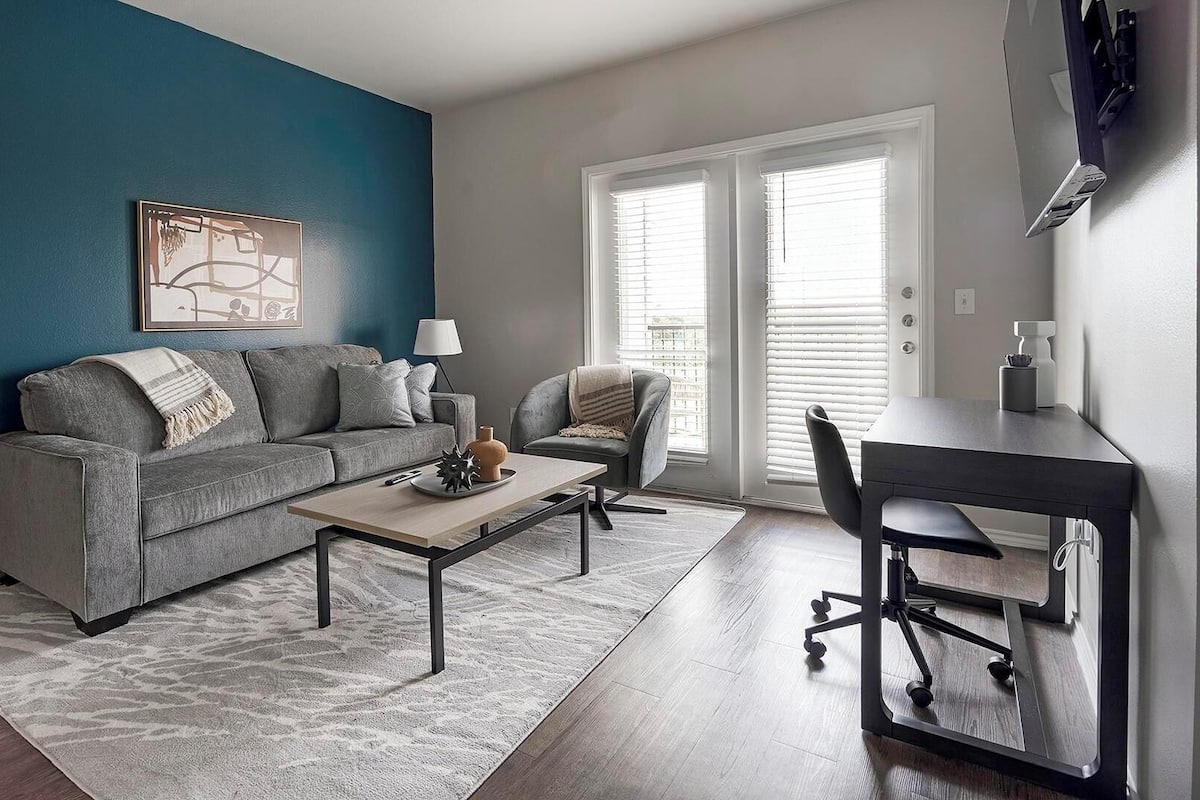 A living room with a gray sofa, a coffee table, a gray armchair, a desk with a chair, and a window with blinds.