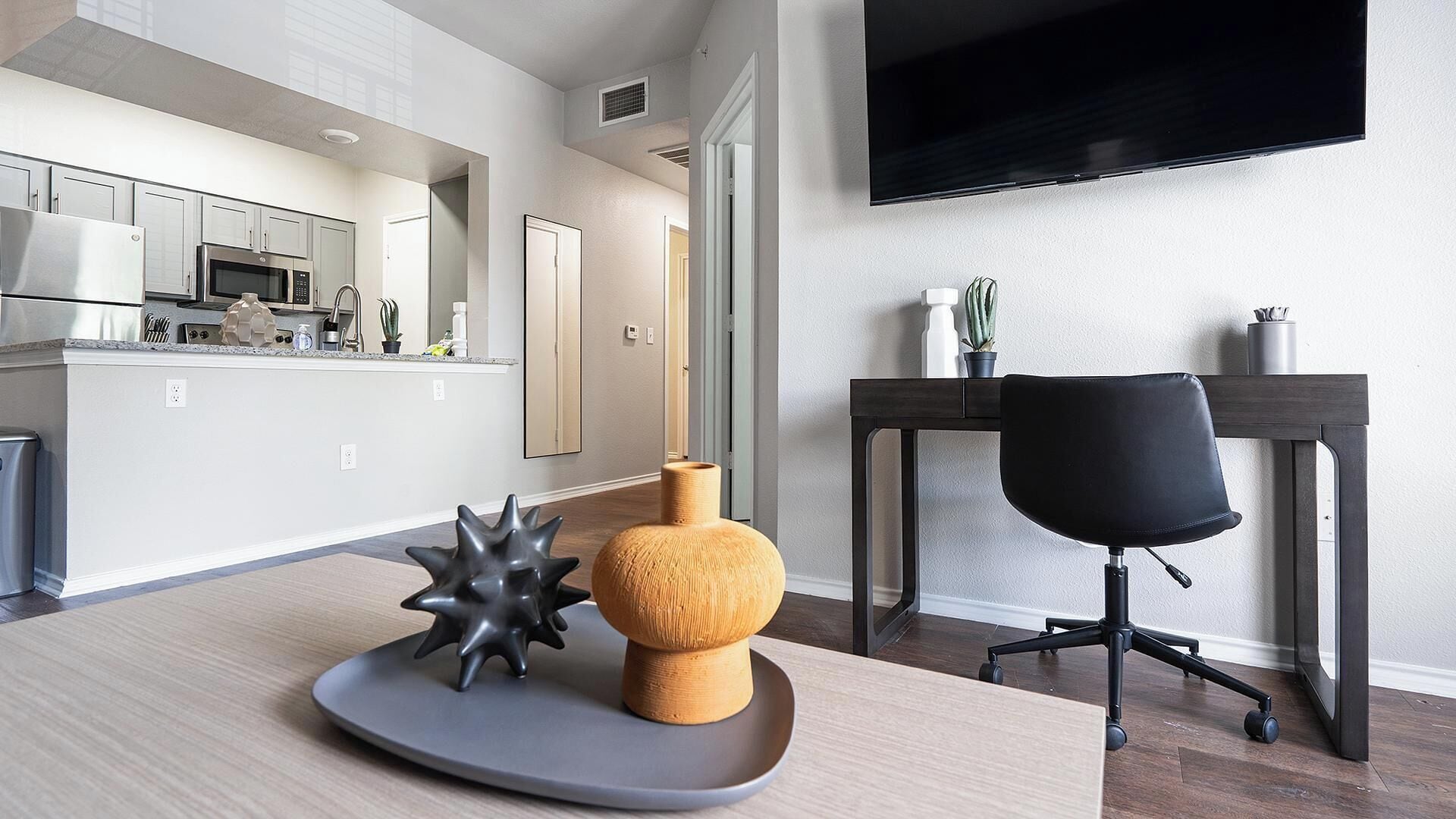 A modern kitchen with stainless steel appliances, a dining area with a wooden table and a black office chair, and a flat-screen TV mounted on the wall.