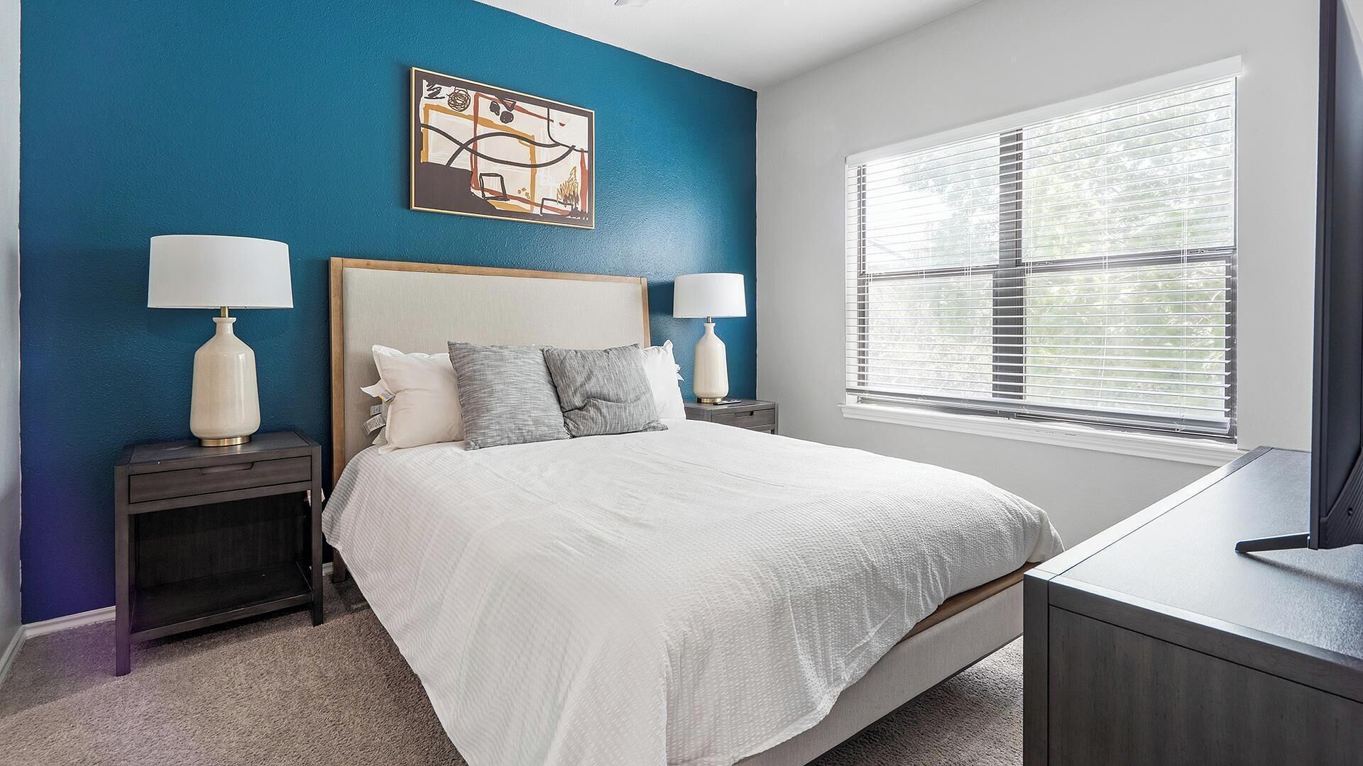 A bedroom with a bed, two bedside tables, a nightstand, a window with blinds, and a wall art piece.