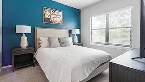 A bedroom with a bed, two bedside tables, a nightstand, a window with blinds, and a wall art piece. - Landing Furnished Apartments - Far West Side (San Antonio)