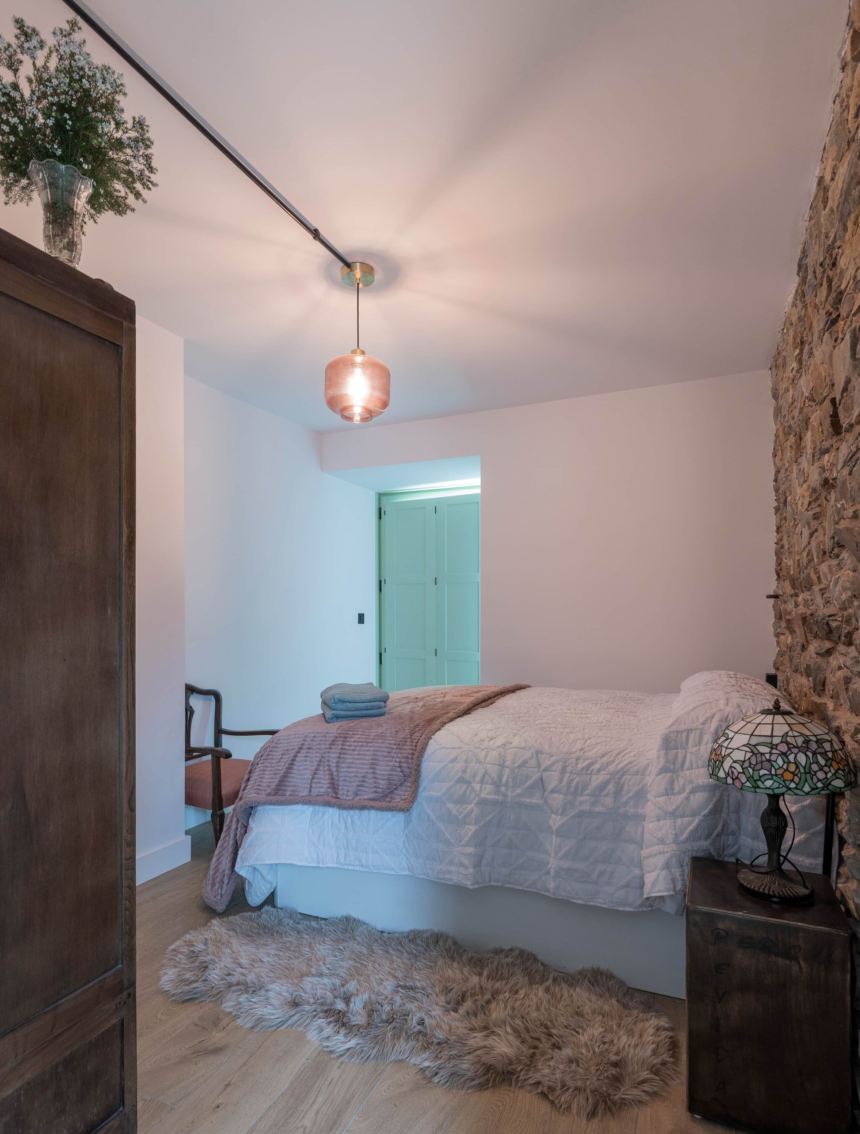 Un dormitorio con una cama, un armario de madera, una pared de piedra, una lámpara colgante y una puerta que da a otra habitación.
