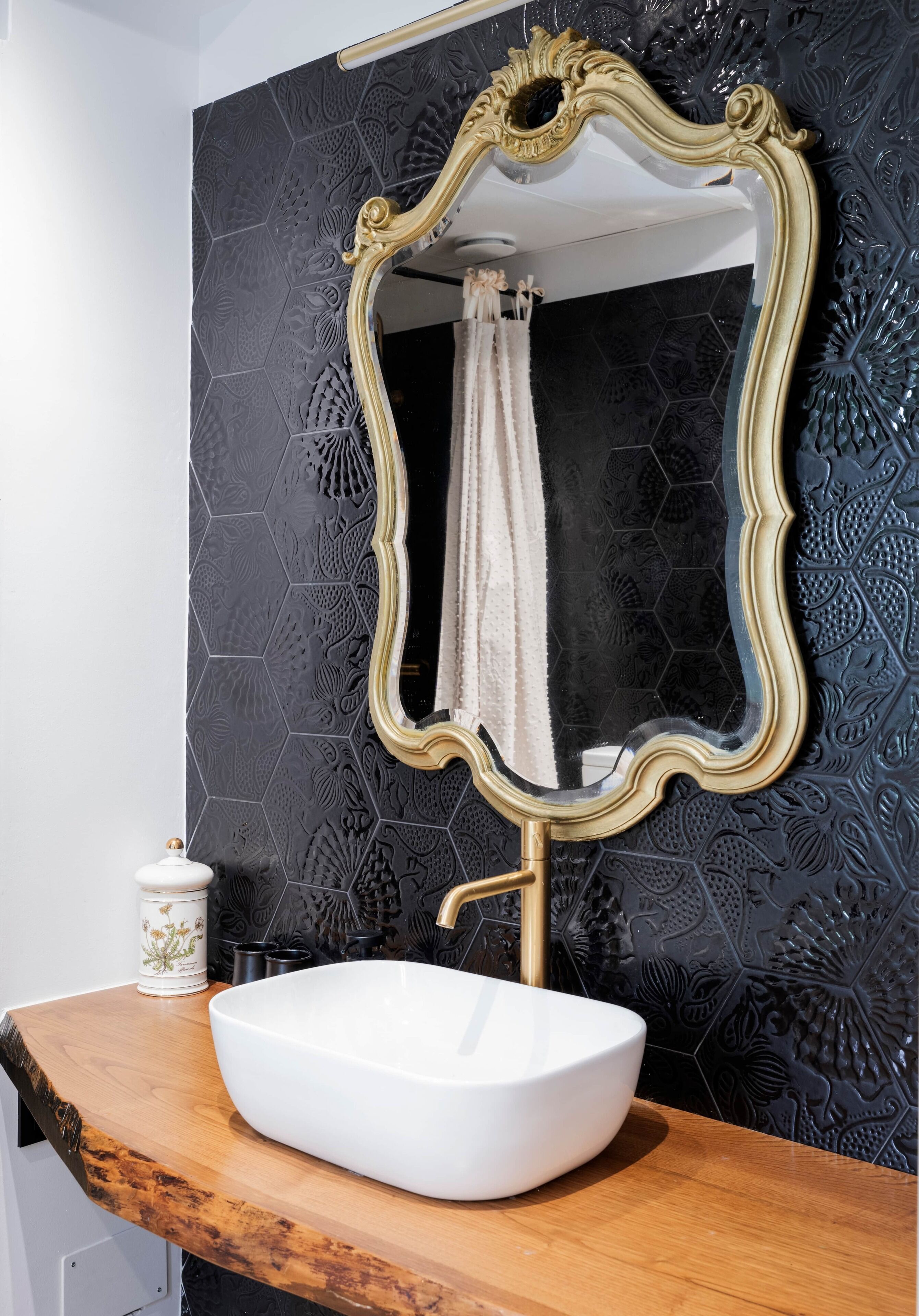Un baño con un espejo grande y decorativo, un lavabo blanco tipo recipiente y una pared con diseño negro.