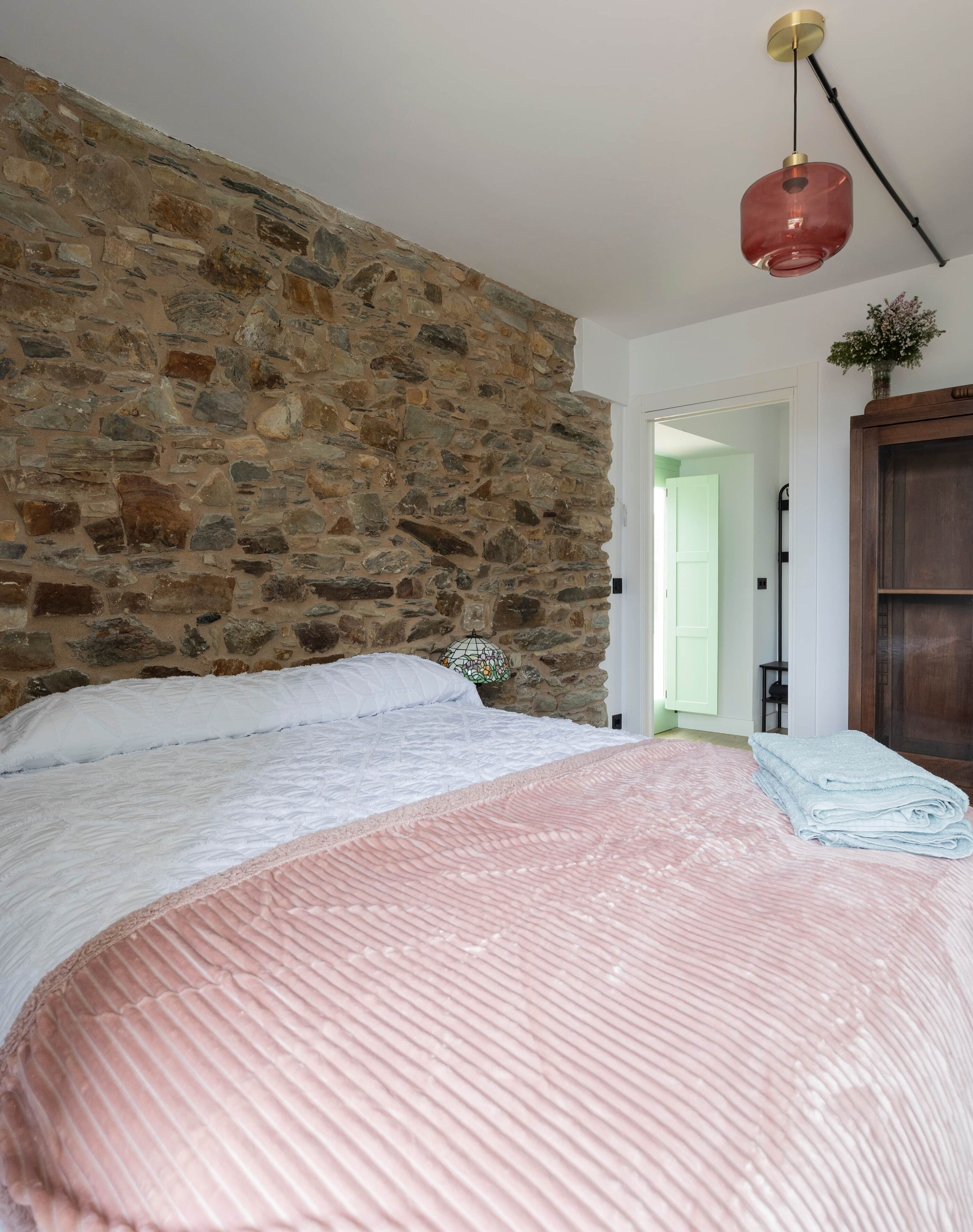 Un dormitorio con pared de piedra, cama con ropa de cama rosa y blanca, lámpara colgante roja y un mueble de madera.