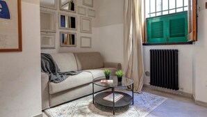 En stue med en beige sofa, et glasbord, en radiator og et vindue med grønne skodder.