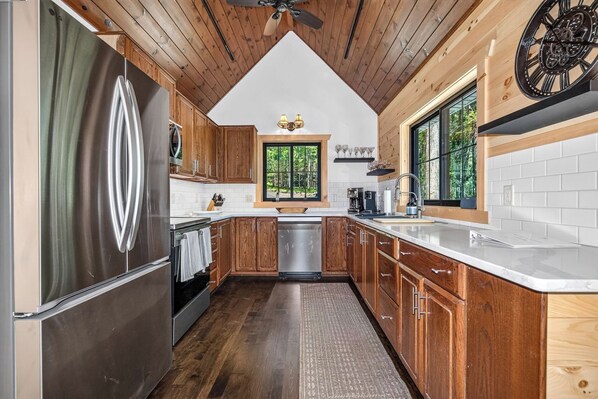 A kitchen with wooden cabinets, a stainless steel refrigerator, a dishwasher, and a window with a view of trees. - An Adventure Awaits -  Stepaway Vacation Rentals (St.Germain)