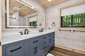 A bathroom with a large mirror, two sinks, grey cabinets, and a window with a view of trees. - An Adventure Awaits -  Stepaway Vacation Rentals (St.Germain)