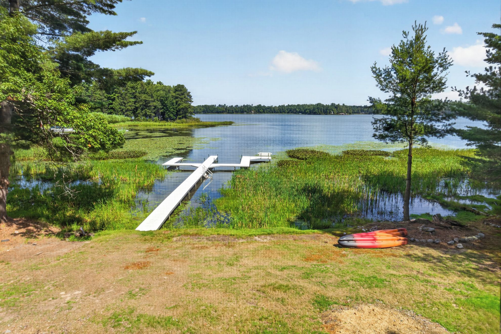 A lakeside dock extending into a calm lake, surrounded by lush greenery and a clear sky.