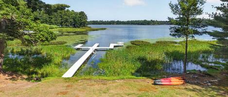 A lakeside dock extending into a calm lake, surrounded by lush greenery and a clear sky.