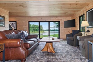 A cozy living room with a brown leather sofa, a wooden coffee table, and a grey armchair. A flat-screen TV is mounted on the wall, and there is a window with a view of a lake. - An Adventure Awaits -  Stepaway Vacation Rentals (St.Germain)