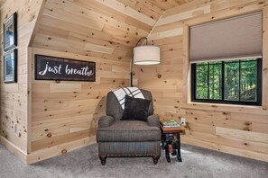 A cozy room with a wooden interior, a comfortable armchair, a lamp, and a window with a view of trees.