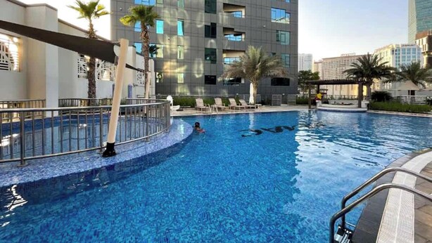 Piscina com uma pessoa nadando, rodeada por palmeiras e um edifício moderno com varandas.