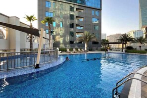A swimming pool with a person swimming, surrounded by palm trees and a modern building with balconies. - StayShort - Stunning Studio Unit in Business Bay that Sleeps 2! (Business Bay)