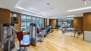 Uma academia moderna com diversos equipamentos de exercício, amplas janelas e piso de madeira.