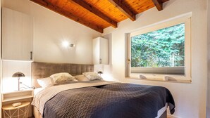 Een slaapkamer met een bed, nachtlampjes, een raam met uitzicht op bomen, en een houten plafond.