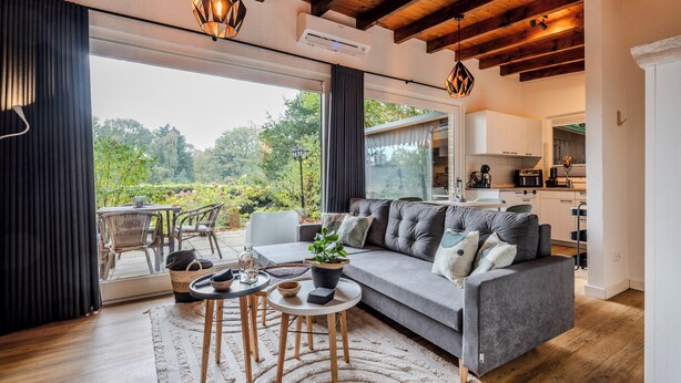 Een moderne woonkamer met een grijze bank, een houten salontafel en uitzicht op de tuin door grote schuifdeuren.
