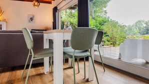 Een moderne eetruimte met een houten tafel en twee stoelen, een grijze bank en een groot raam met uitzicht op groen.