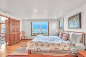 A bedroom with a large bed, a view of the ocean through a window, a wooden door, and a chair. - Wileys Wavehouse: Oceanfront Yachats Retreat Near Cape Perpetua (Yachats)