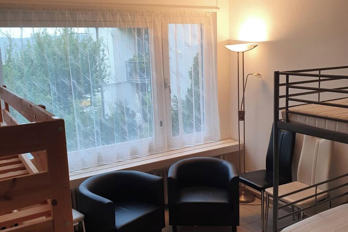 Une chambre avec un lit superposé, deux chaises, un bureau, une lampe et une fenêtre donnant sur des arbres.