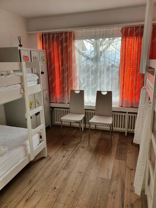 A room with bunk beds, two chairs, a radiator, and a window with red curtains.