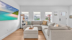 En rummelig stue med en stor hjørnesofa, en pudeskammel og et maleri af en strandscene. Tre vinduer med skodder, en dør med et skilt og en lampe på et sofabord.